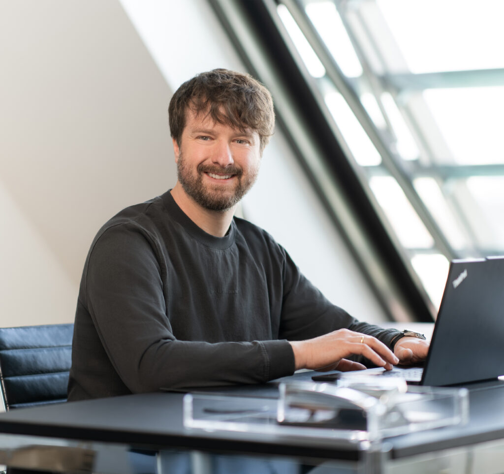 Porträt eines Mitarbeiters der kaufmännischen Abteilung, lächelnd am Laptop in einem hellen Büro – BLOCK Immobilienmanagement
